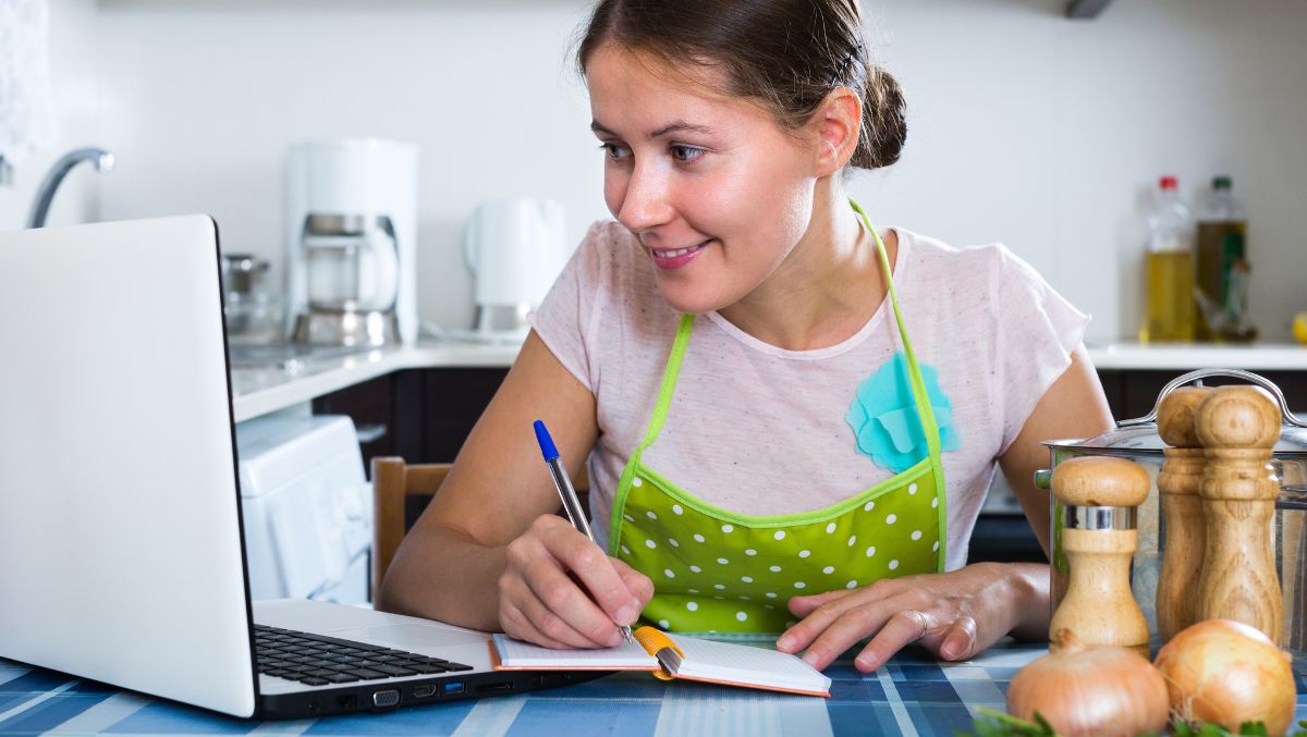 Woman c5reating grocery list on laptop at kitchen counter